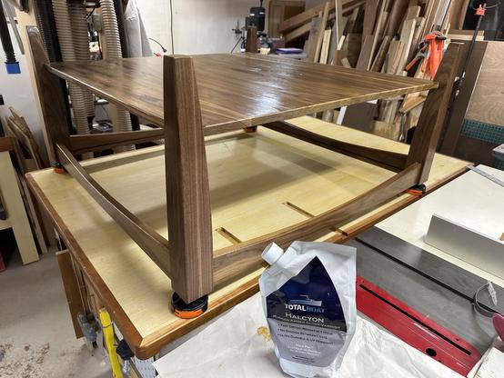 A wooden table with a large, flat surface supported by four legs is shown in a workshop. Nearby, there is a pouch of TotalBoat Halcyon varnish placed on a work surface. Woodworking tools and materials are visible in the background.
