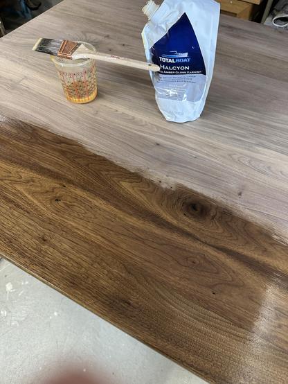 A tabletop made of wood, with one area finished and another area awaiting treatment. A paintbrush rests in a measuring cup containing a gloss varnish labeled "TotalBoat Halcyon," positioned next to a pouch of the same varnish.