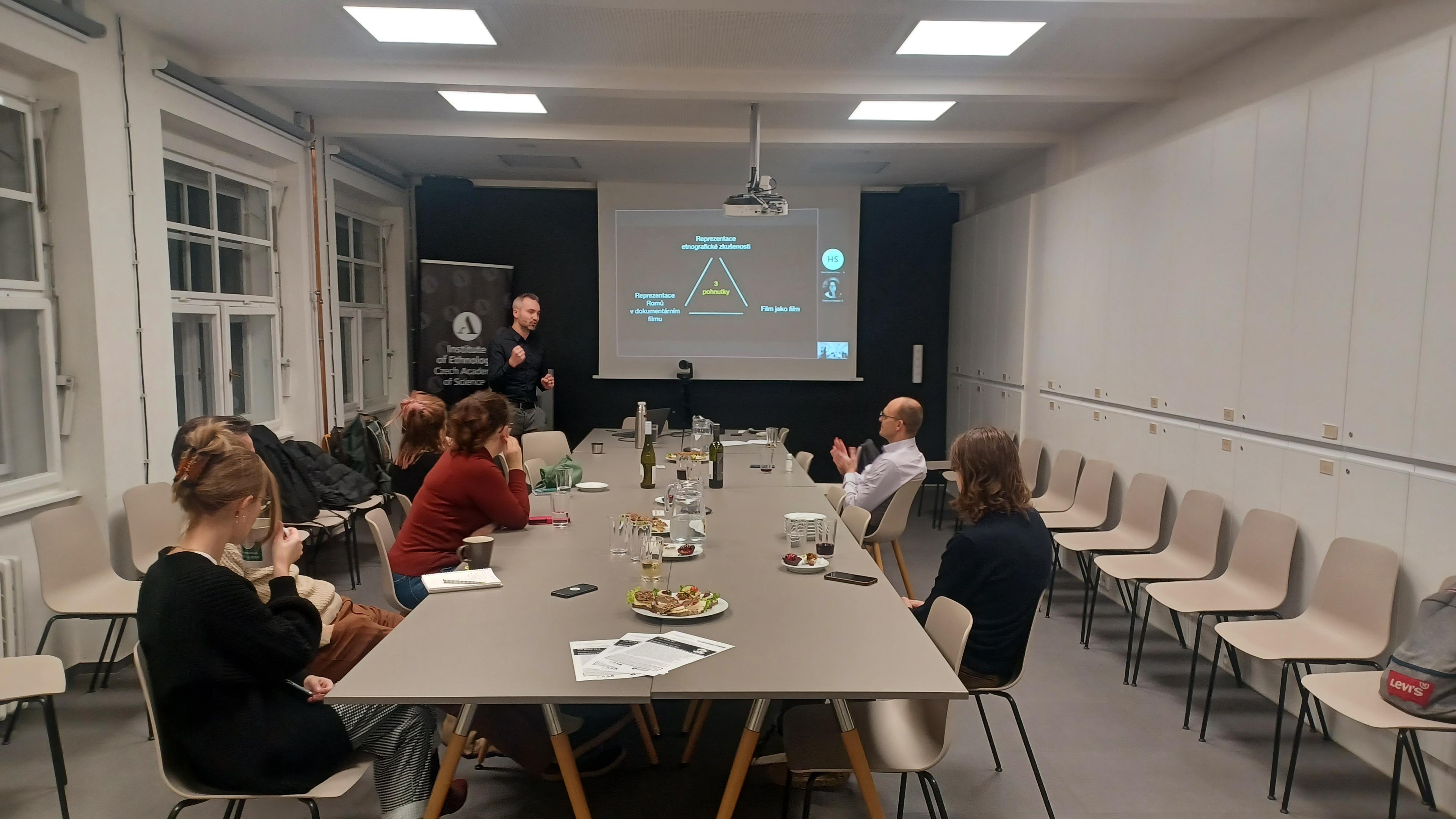 6 people + 1 speaker having a seminar in a seminar room, there's a screen in the background.