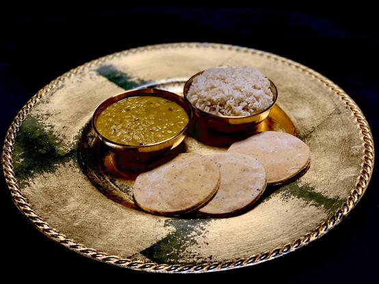 On a gold-looking plate are two gold looking bowls with three baked large round crackers arranged neatly on the plate. In the two bowls are a dark yellow curry tinged green from leaf powder and a rounded bowl of rice. The background is black. 