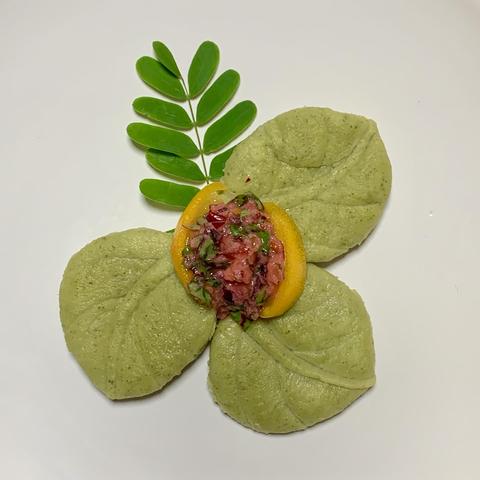 White dish with 4 leaves pointing in every direction. The top leaf is a dark green tamarind leaf, the other three leaves are jackfruit pasta molded into a leaf with a mooncake mold. In the center is two thin slices of Fukushu kumquat that hold together the dressing which is piled on top. The kumquat is light orange and the dressing is dark pink with bits of red veined green chopped leaves. 
