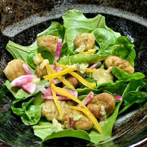 In a black decorative bowl with white stripes is a colorful salad with pink and orange strips and green leaves. There are small fried gnocchi on the leaves that if you look past the Maillard-reaction, you can see they’re green. There are drops of a light yellow salad dressing. 