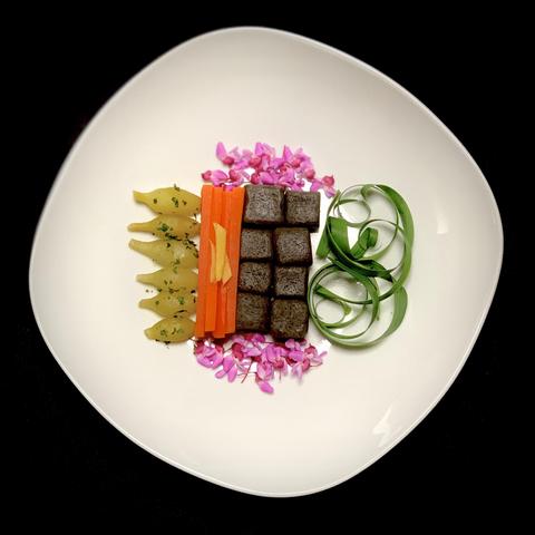 A white plate featuring a decorative arrangement of food, including dark square pieces, sliced carrots, green vegetables, pale yellow pieces, and pink flower petals. The presentation is artistic and colorful against a black background.

The native plants are redbud flowers and southern live oak dumplings. 