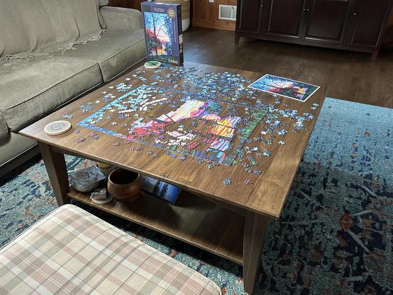 A wooden coffee table displays a partially completed 1000-piece puzzle, with scattered pieces around it. Next to the puzzle, there's a printed image of the completed puzzle and a coaster. A plaid-patterned cushion is visible beside the table.