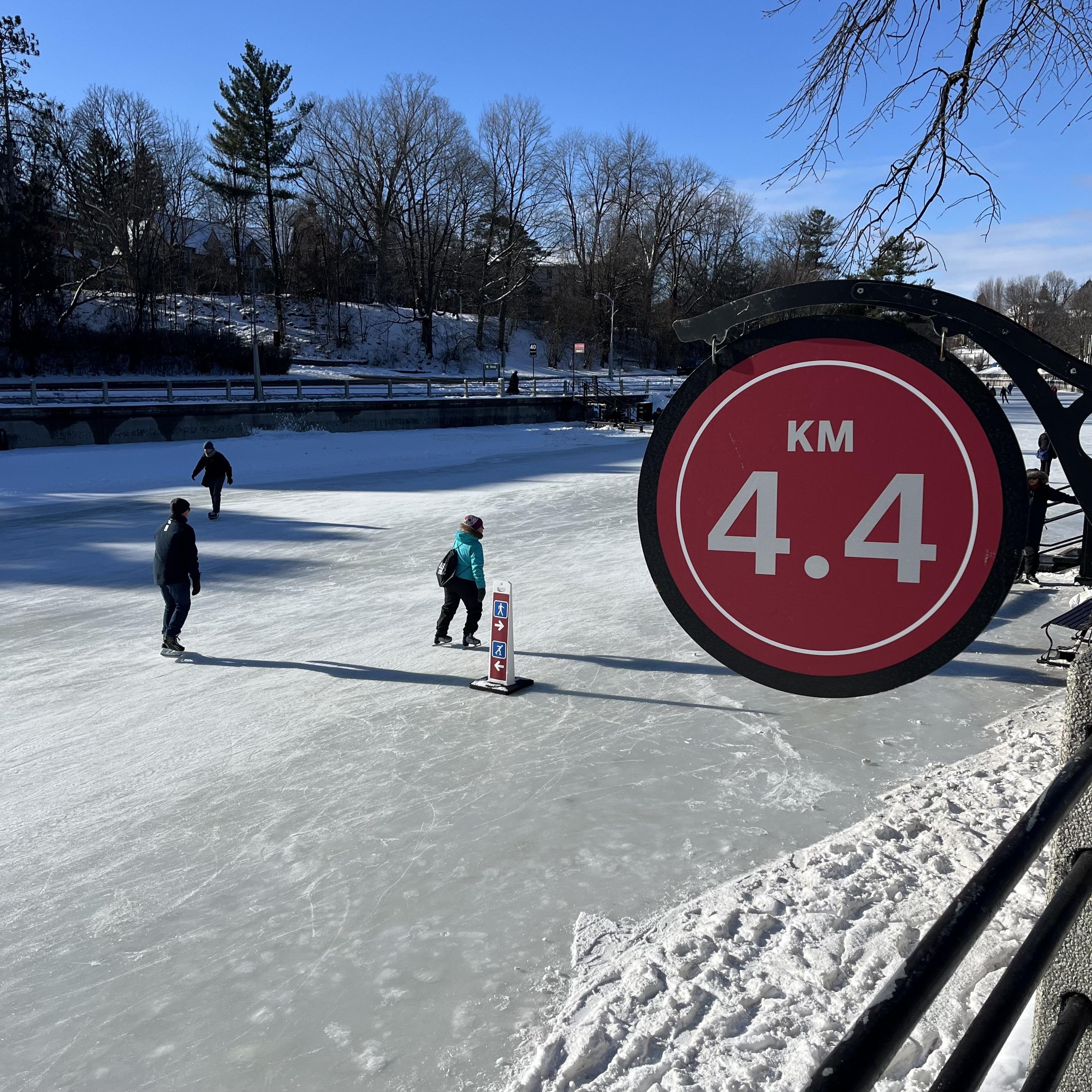 A frozen pathway with ice skaters gliding on it. A prominent sign displays "4.4 KM.&quot; Snow-covered banks and trees are visible in the background under a clear blue sky.