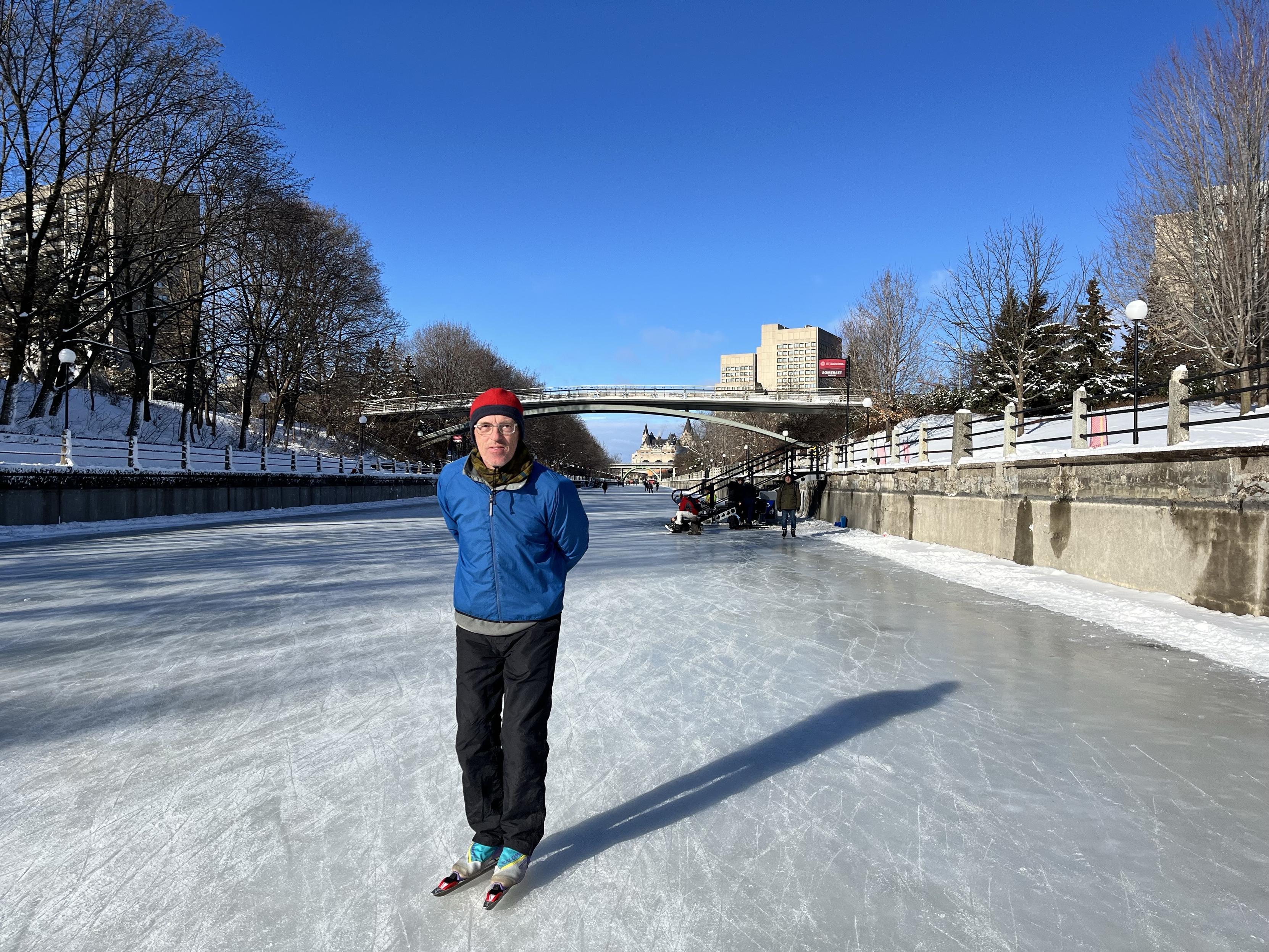 A man stands on ice while wearing a blue jacket, black pants, and a red hat. The scene features a winter landscape with snowy trees and buildings in the background, under a clear blue sky. There are people in the distance on the ice