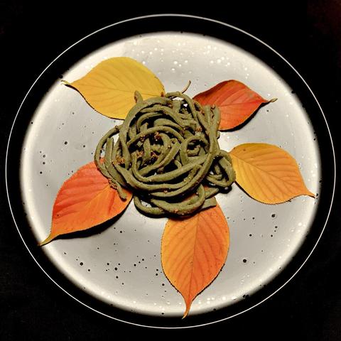 A plate features a twisted green pasta arranged in a circular shape at the center, surrounded by vibrant autumn leaves in shades of orange and yellow. The background is dark, enhancing the colors of the leaves and food.

On the pasta is popped huazontle grains. 