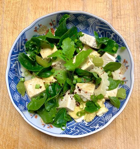 A vibrant salad featuring cubes of tofu, mixed greens, and chopped green madhatter peppers, white bitter melons, epazote, garnished with a sprinkle of red pepper flakes. The dish is presented in a decorative blue and white bowl, placed on a wooden surface.