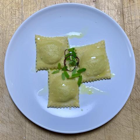 A white plate with three pieces of ravioli, drizzled with oil and garnished with small green tamarind leaves and a sprig of red garnish (peeled red veined sorrel stem). The background shows a wooden surface.