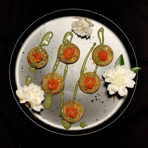 A round black plate with six circular bites, each topped with a dollop of red sauce and green drizzles, is surrounded by white flowers. The background is dark. 

Mini acorn okonomiyaki with madhatter/queen of minalco tomatillo/strawberry and yuzu sauce, sissoo-ezu and yuzu zest. Adapted from “Okonomiyaki.“ Sakai, p.261. 