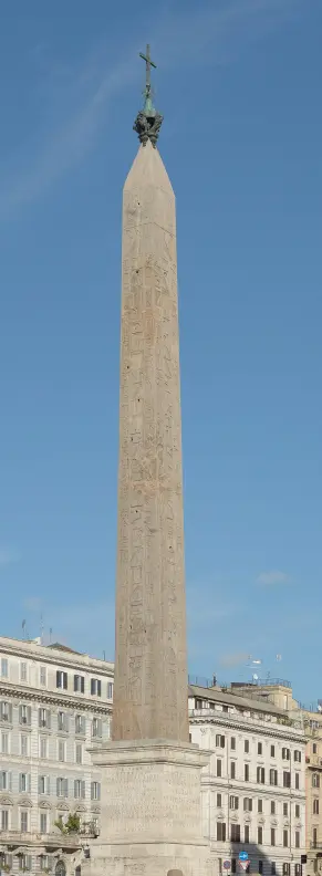 The image depicts an ancient obelisk. Obelisks are tall, four-sided, narrow tapering monuments with a pyramidion on top, often covered in hieroglyphs. This specific obelisk is located in a public square, surrounded by buildings and features a cross at its apex, indicating it may have religious significance. The surroundings include a mix of architectural styles typical of urban settings.

Image Credits: Wikimedia / Livioandronico2013 / CC BY-SA 4.0