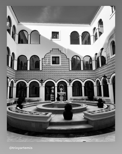 A black and white photo of a courtyard there is a circular fountain at the centre of the courtyard with circular seating and flower beds around it. The building is in a quad and has small arches all the way round in a quad and is three stories high with ornate windows with a Moroccan influence in style. The window reflect the opposite windows and it feels like reflection upon reflection. There is a nice sharp geometric shard of light from the sun on the top right of the frame. 
