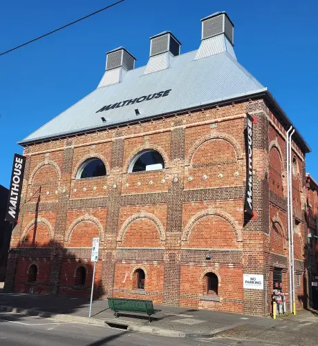 The image shows a brick building with a distinct architectural style. The building has a peaked roof with multiple vents and large arched windows. There are signs indicating "Malthouse" on the structure. The foreground includes a bench and a sign indicating "No parking." The weather appears clear, with a blue sky in the background.

Image Credits: Wikimedia / Shkuru Afshar / CC BY-SA 4.0