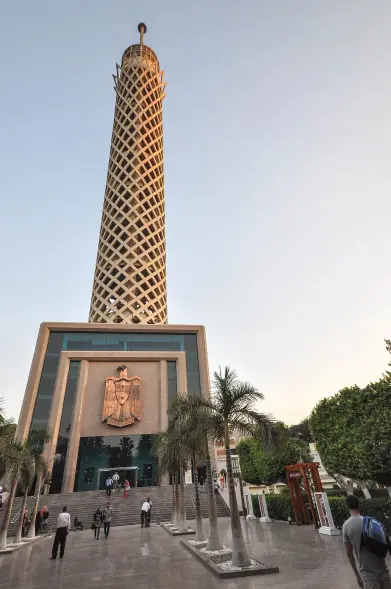 The image depicts a tall tower with a distinctive design, likely set in a landscaped area with palm trees. The tower appears to have a lattice-like structure and is accompanied by a building at its base that features a carved emblem or symbol. People can be seen walking in the vicinity, indicating it may be a public attraction or landmark. The setting suggests it could be in a city with a warm climate, possibly during sunset or sunrise.

Image Credits: Wikimedia / Jorge Láscar from Melbourne, Australia / CC BY 2.0