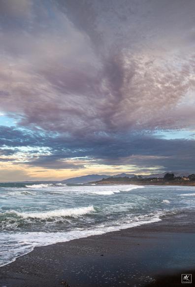 Color photo of waves breaking on a beach under partly cloudy early morning skies. 