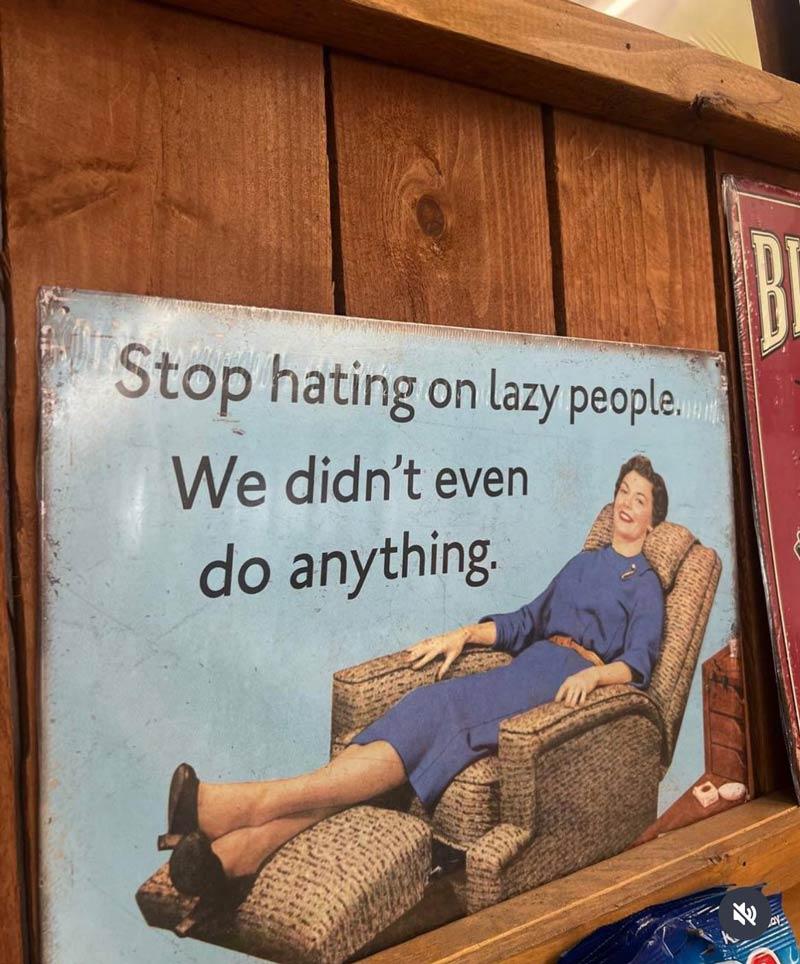 Humorous tin sign reading, "Stop hating on lazy people. We didn't even do anything.&quot; The sign features a vintage advertisement of a woman relaxing in a chair with her feet up.