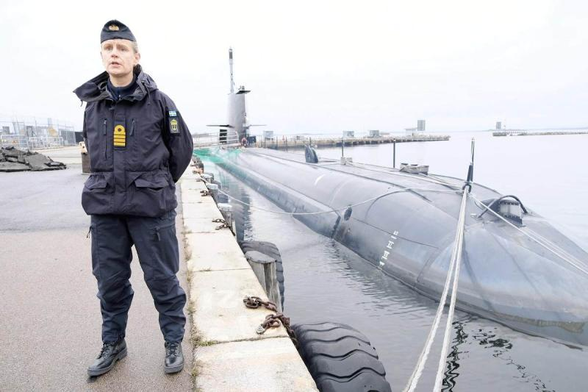 Paula Wallenburg, Swedish submarine commander, in a navy uniform stands on a dock next to a large submarine. 

Paula is wearing a dark blue jacket with several patches and a cap. The submarine is moored to the dock with ropes and is partially submerged in the water. 

The dock is made of concrete, and there are large black fenders attached to it.

In the background, there is a calm body of water and a distant shoreline with some buildings visible. The sky is overcast, giving the scene a muted, gray appearance.