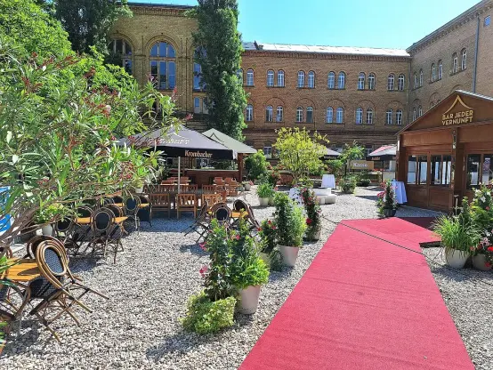 The image depicts a charming outdoor space, likely a café or restaurant courtyard. There are tables and chairs set up under umbrellas, with a red carpet leading through the area. Numerous potted plants and flowers add color and greenery, creating a pleasant atmosphere. The background features a large brick building with windows, suggesting an inviting and communal setting.

Image Credits: Wikimedia / Fridolin freudenfett / CC BY-SA 4.0