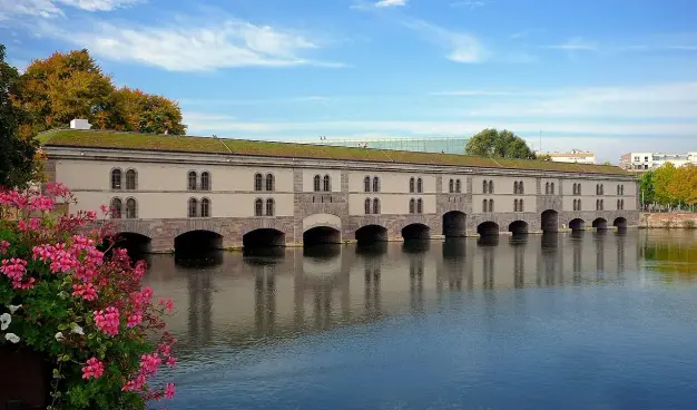 The image shows a large building situated near a body of water. The structure has multiple arched openings, a green roof, and is reflected in the water. Surrounding the building, there are flowers in the foreground and trees in the background, with a clear blue sky above. This scene conveys a tranquil, picturesque setting.

Image Credits: Wikimedia / Claude TRUONG-NGOC / CC BY-SA 3.0