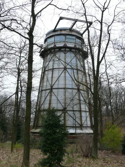 The image shows an old, tall structure that resembles a water tower or a lighthouse. It has a cylindrical shape with a glass or transparent upper portion, surrounded by a metal framework. The tower is located in a wooded area, with several trees nearby. The scene appears to be somewhat overcast or gloomy, indicating that it may be a cooler season.

Image Credits: Wikimedia / brandenburgref / Public domain