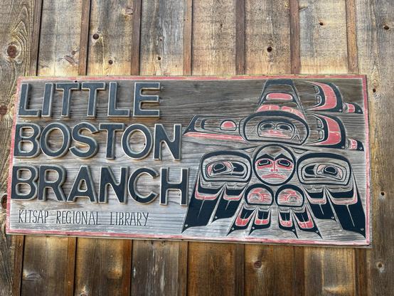 A wooden sign for the Little Boston Branch of Kitsap Regional Library features stylized indigenous artwork. The design includes faces and figures in black and red, set against a wooden background.