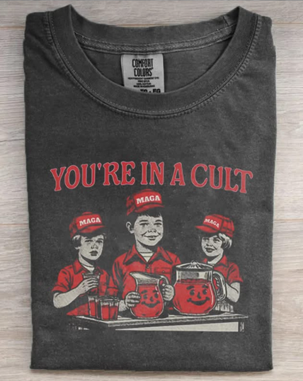 The image shows a dark gray t-shirt with a graphic design printed on the front. The design features three individuals wearing red caps with the text "MAGA" on them, and red shirts. They are depicted behind a table with two large pitchers and several glasses, all filled with a red liquid. The pitchers have smiling faces on them. Above the individuals, the text "YOU'RE IN A CULT" is printed in bold red letters.
