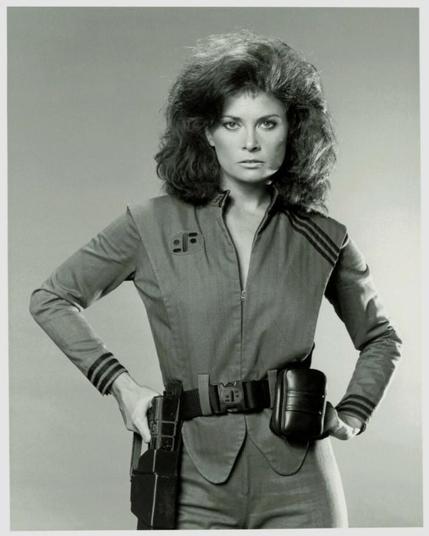 Pla americà de Diana (actriu Jane Badler) de la clàssica sèrie de televisió "V" dels anys 80. Mira la càmera amb expectació mentres treu la seva pistola que penja del cinturó. Va vestida amb l'uniforme dels "Visitants", la cremallera baixada a mitja alçada. / Medium shot of Diana (actress Jane Badler) from the classic TV series "V" of the 80s. She's looking at the camera with expectation as she pulls the gun hanging from her belt. Dressed in "Visitant" uniform  the zipper has been pulled down half way through.