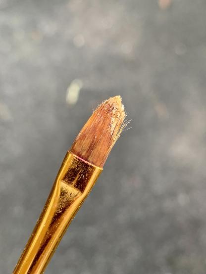 A close-up of a paintbrush with a gold handle and orange bristles coated and smattered with yellow pollen. The background is blurred and dark, focusing on the brush.