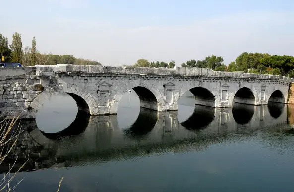 The image depicts a stone bridge with multiple arches spanning over a body of water. The bridge appears to be historic, constructed from large stone blocks. Surrounding the bridge are trees and a clear sky, suggesting a serene landscape. The water beneath reflects the bridge, enhancing the picturesque scene.

Image Credits: Wikimedia / Heiko Trurnit / CC BY-SA 2.5