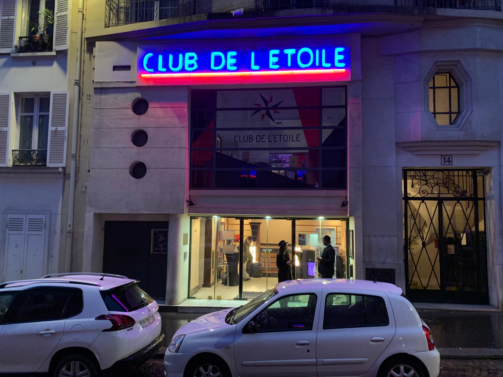 Evening scene, two silhouettes of people hanging in from of a cinema. A big neon letters read "Club de L'etoile". Photo by Michael Brims.