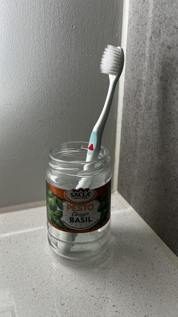 A toothbrush is placed upright inside an empty glass jar labeled "Pesto Classic Basil." The jar is sitting on a light-colored countertop. The background consists of textured wall surfaces.