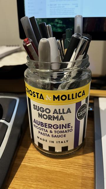 A glass jar labeled "Sugo alla Norma" pasta sauce containing various pens and markers is placed on a wooden surface, with a blurred computer monitor in the background.