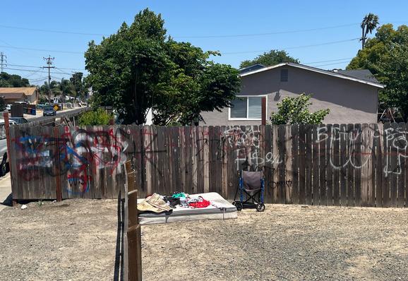 A bare mattress topped with scattered clothing in a dirt median next to graffiti'd wooden fence behind which is a house. There is a wheelchair next to the mattress.