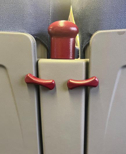 A red pommel between a pair of seat backs, below which are a pair of red wishbone shaped clips to hold up the fold-out seat back trays, all on a beige background. The red pommel and the two cups resemble a head a forearms held in the manner of a bodybuilder doing a double flex.