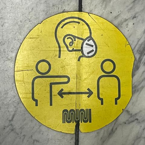 A floor sticker encourages mask wearing and social distancing on Muni vehicles. The top is a line drawing for a man's head in profile wearing a mask, and the bottom are two line figures, akin to stick figures, with one with its arm out toward the other and and a two-headed arrow indicating the proper distance.