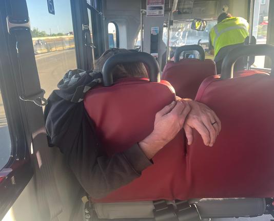 Seen from behind, a bus passenger hugging his seat, with one arm going around and the other sliding past the gap between his seat and the next, the hands touching, his head down. 