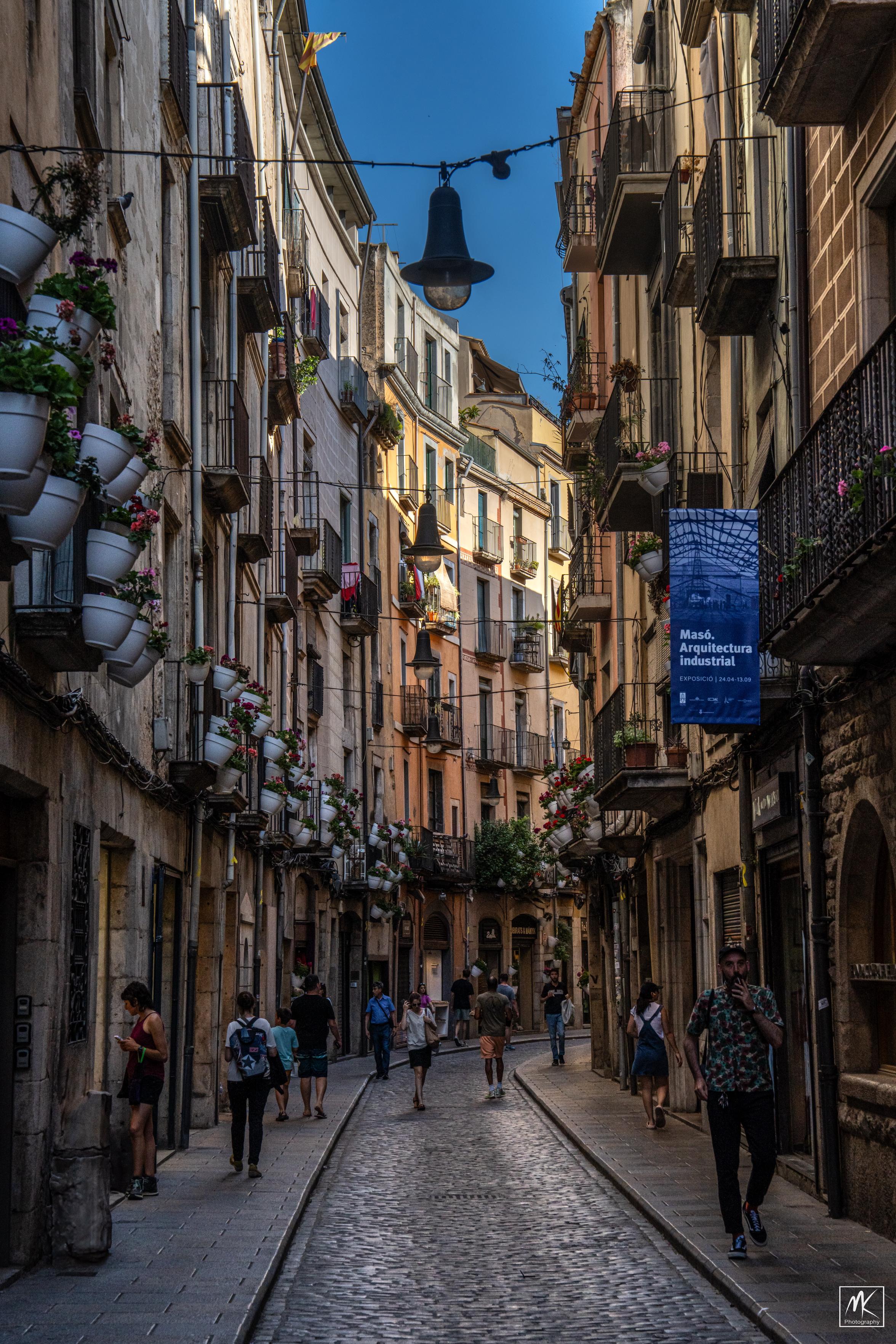 Color photo of a narrow, curving street with several pedestrians in Girona, Spain. 