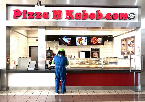 A man with a green cap and blue coveralls ordering at a Pizza N Kabob