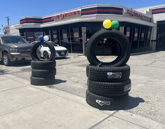 Two sets of tires stacked up, each with another tire "standing up" on top with a pair of cantaloupe-sized balloons on top, roughly reminiscent of Mickey Mouse ears, outside a tire dealership 