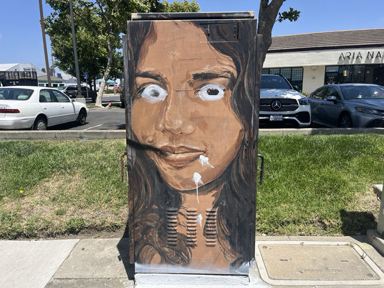 A painting of a crazy-eyed young woman with white drips below her mouth on a utility box in front of a strip mall 