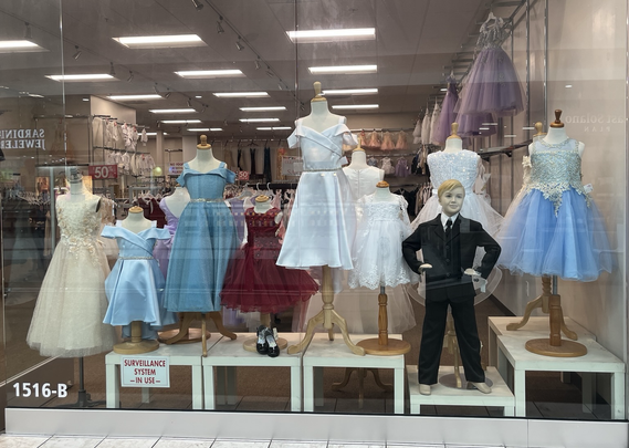 A shop window with eight gowns on dress forms, in front of which is a boy mannequin a natty little business suit with his hands on his hips