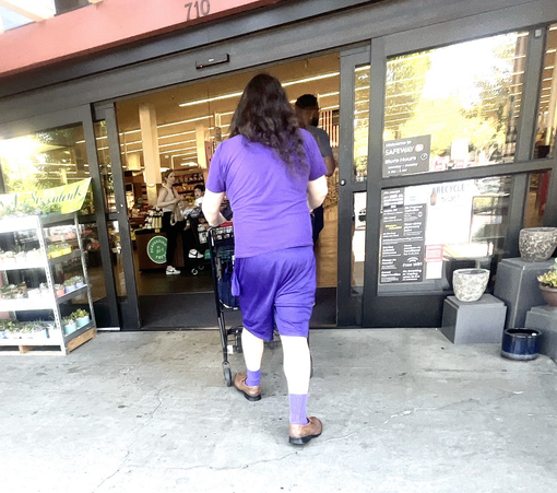 Seen from behind, a long-haired man entering a supermarket. He wears a purple T-shirt, long purple shorts, purple socks, and, proving he's no slave to fashion, brown shoes.
