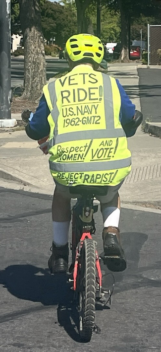 A man on a bike wearing a yellow visibility vest, seen from behind. The vest reads VETS RIDE!  U.S. NAVY 1962-GMT2 RESPECT WOMEN! AND VOTE! REJECT THE RAPIST!  