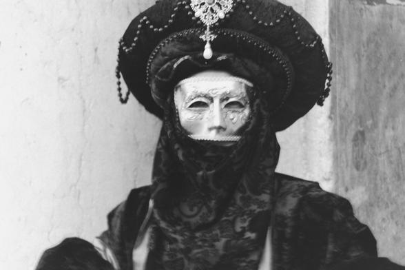 The image features a person dressed in elaborate Venetian-style attire, likely from a historical or cultural event. The individual is wearing a large, ornate hat adorned with decorative elements, including a prominent brooch and dangling beads. The hat is dark, possibly black, with a wide brim and intricate detailing. The person is also wearing a black, patterned veil that covers the head and neck, with a lace-like design. The veil is draped over the shoulders, adding to the dramatic effect of the costume. The background is a plain, light-colored wall, providing a stark contrast to the dark attire. The overall composition is monochromatic, emphasizing the textures and details of the costume.

Provided by @altbot, generated privately and locally using Ovis2-8B

🌱 Energy used: 0.173 Wh