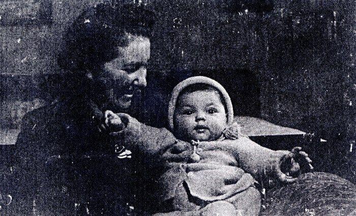 Old grainy black and white photo of a woman smiling at a baby who is sitting up and looking slightly to the side