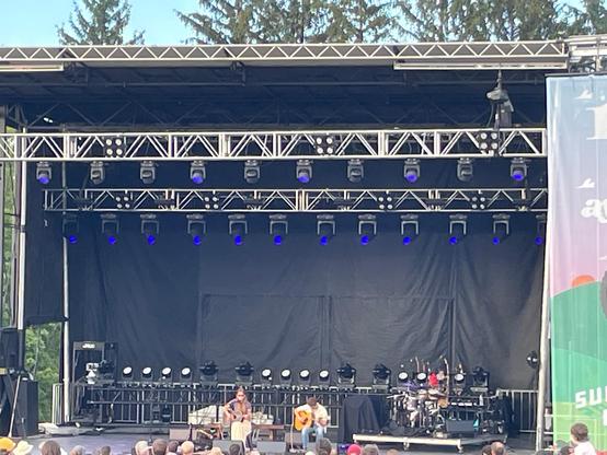 Two performers with guitars sit on an outdoor stage.