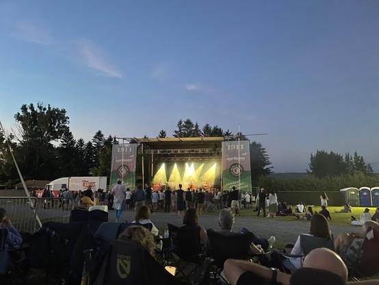 An outdoor stage lit up with yellow lights. There are concert-goers seated and standing around the stage.