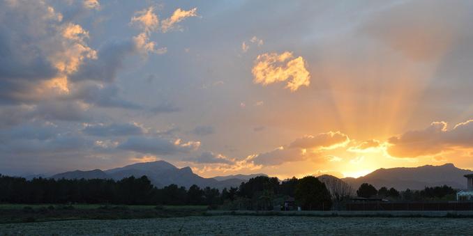 A photo of the sun setting behind distant mountains. There's a field in the foreground. The sky is cloudy. There are crepuscular rays coming over the mountains.