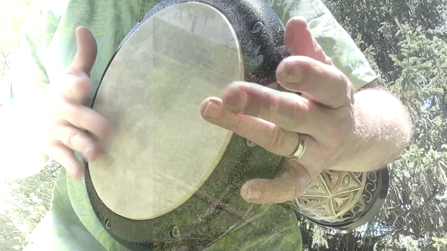 The video features a person playing a djembe drum outdoors, wearing a light green shirt and a gold ring on their left hand. The drum, with a black frame and a light-colored skin, is held close to the camera, highlighting the intricate patterns on its side. The person's hands are actively engaged in playing the drum, with fingers and palms striking the drumhead in various rhythmic patterns. The background is a natural setting with trees and sunlight filtering through the leaves, creating a seren…