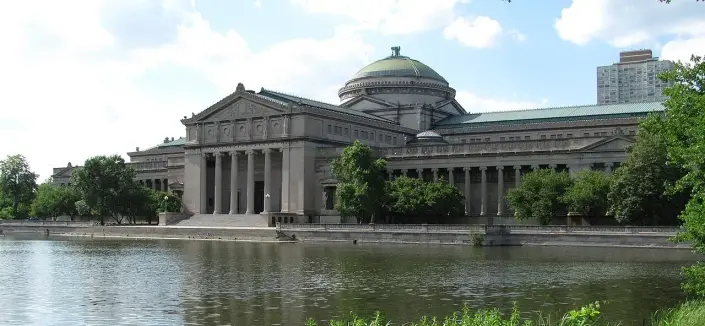 The image shows a prominent building with a large domed roof and classical architectural features, such as columns and a grand entrance. It is situated near a body of water, likely a lake or river, and is surrounded by greenery. The setting suggests a public building, possibly a museum or similar institution.

Image Credits: Wikimedia / Knarfol at de.wikipedia / Public domain